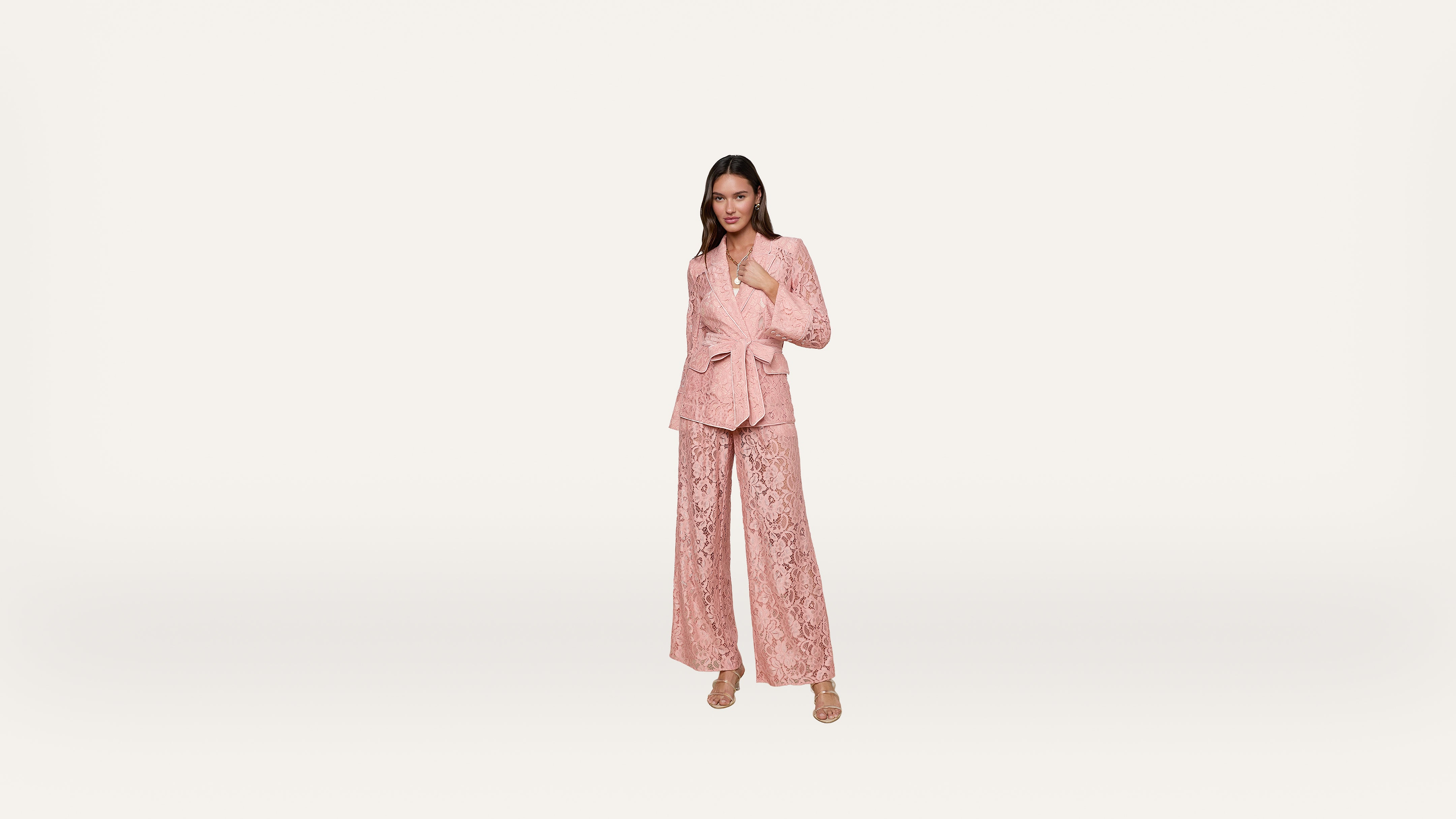 Woman standing and smiling, wearing a light pink lace suit with wide-leg pants and matching blazer, and beige open-toe heels, against a plain white background.