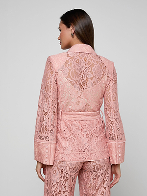 A woman faces away, wearing the Jordana Lace Blazer in light pink. The sheer floral lace design with long sleeves and a belted waist reveals matching undergarments, perfect for Spring 2026 against a plain, light-colored background.