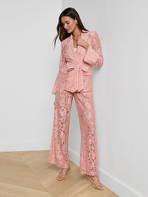 A woman indoors wears the Jordana Lace Blazer—a stylish, light pink sheer floral lace pantsuit with flared pants and a belted evening jacket. With long brown hair, she leans against a white wall for a chic Spring 2026 look.