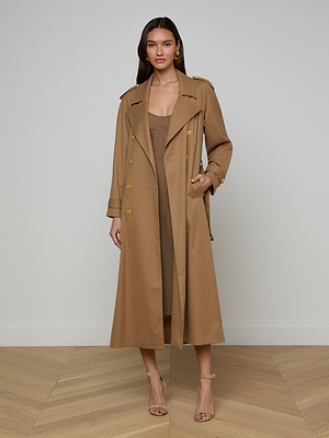 A woman with long brown hair stands in a minimalist room, facing forward and smiling slightly. She wears the Brinley Oversized Trench Coat belted at the waist over a matching dress and strappy beige heels.