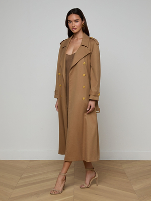 A woman with long brown hair stands indoors by a plain wall, wearing the Brinley Oversized Trench Coat with gold buttons over a tan dress, beige heeled sandals, and gold earrings.