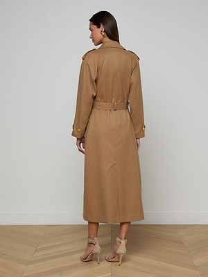 A woman stands indoors facing away in the Brinley Oversized Trench Coat with a belted waist and buttoned cuffs, styled with nude high-heeled sandals and green earrings, on a wooden floor before a white wall.