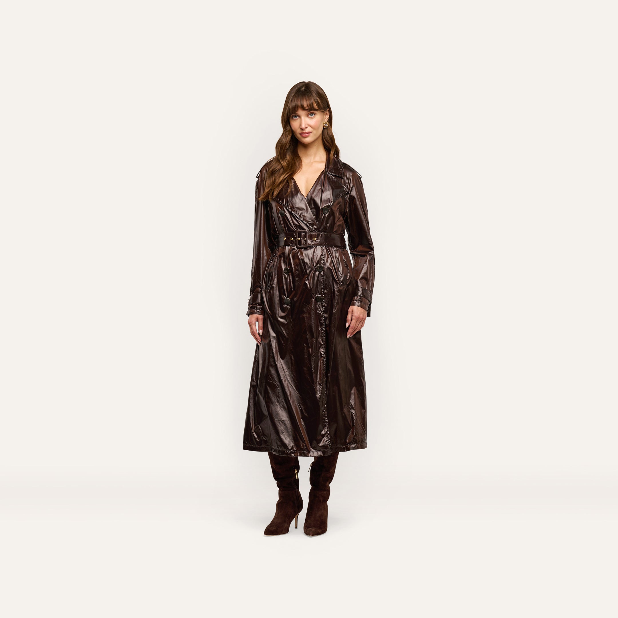 A woman wears an oversized, double-breasted Brinley Nylon Trench Coat in a dark, glossy finish, paired with dark brown heeled boots.