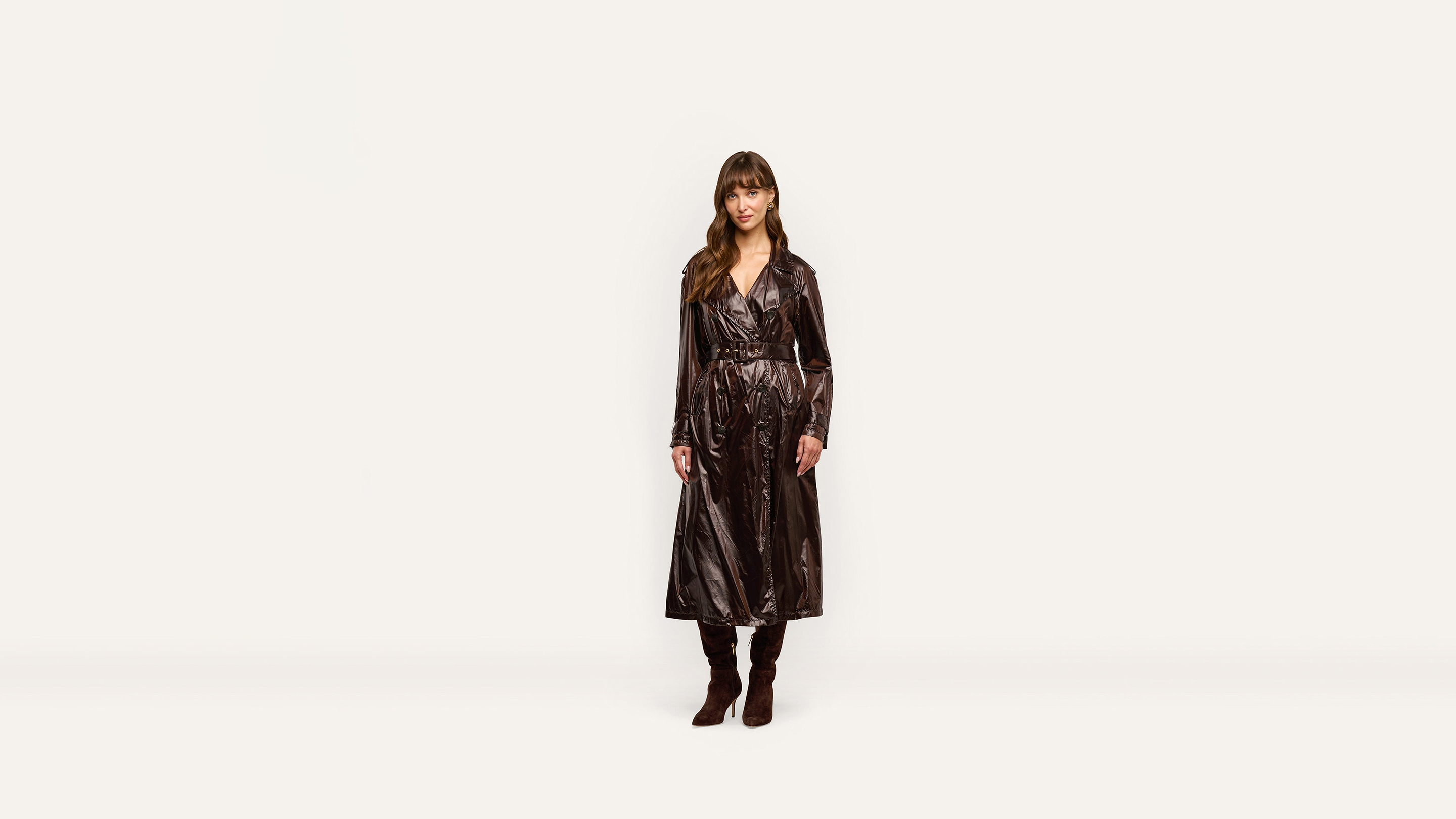 A person wearing a long, glossy belted trench coat and brown knee-high boots stands against a white background.