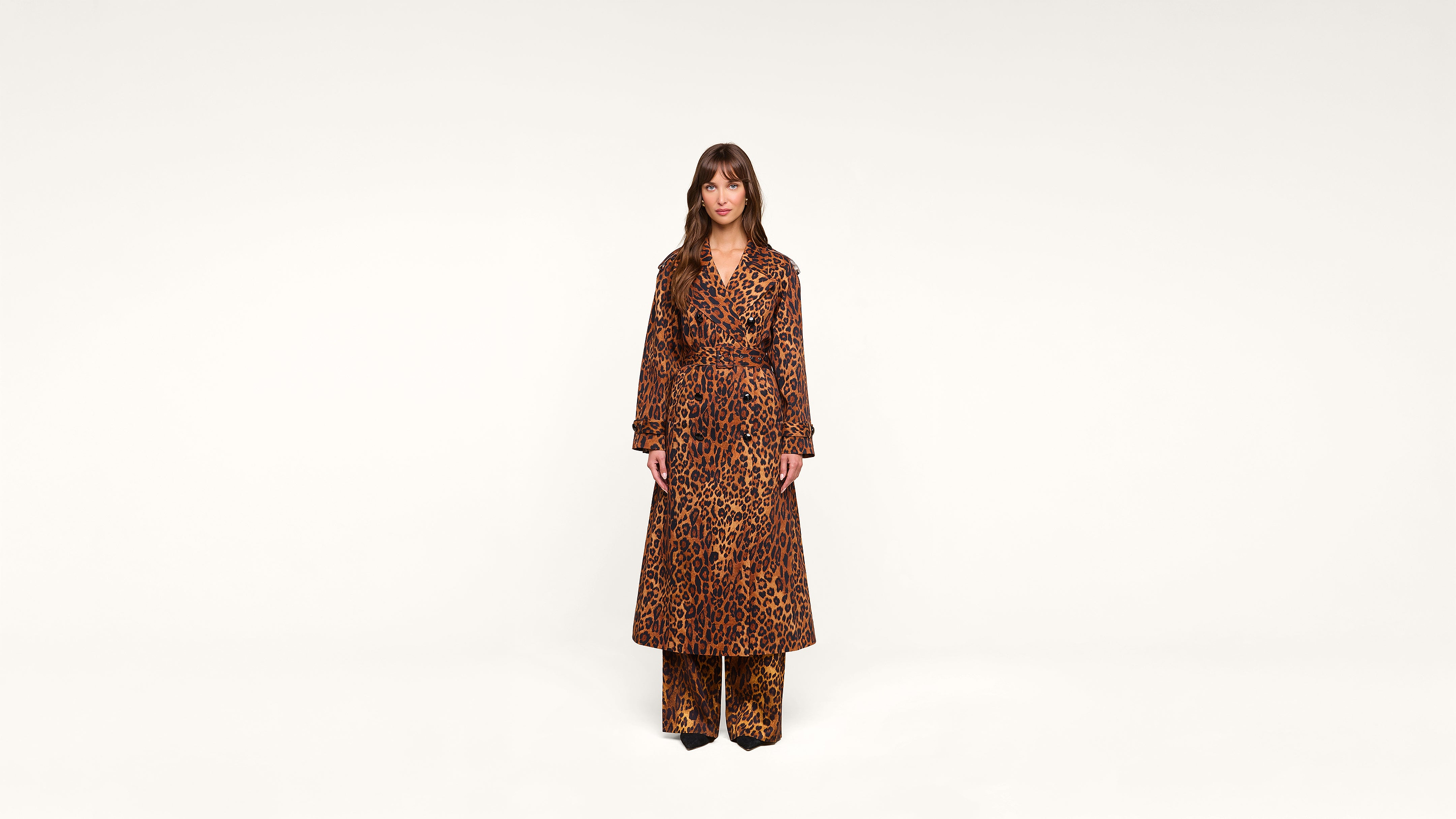 A woman stands against a plain white background, wearing a long leopard-print coat over matching leopard-print pants and black shoes.