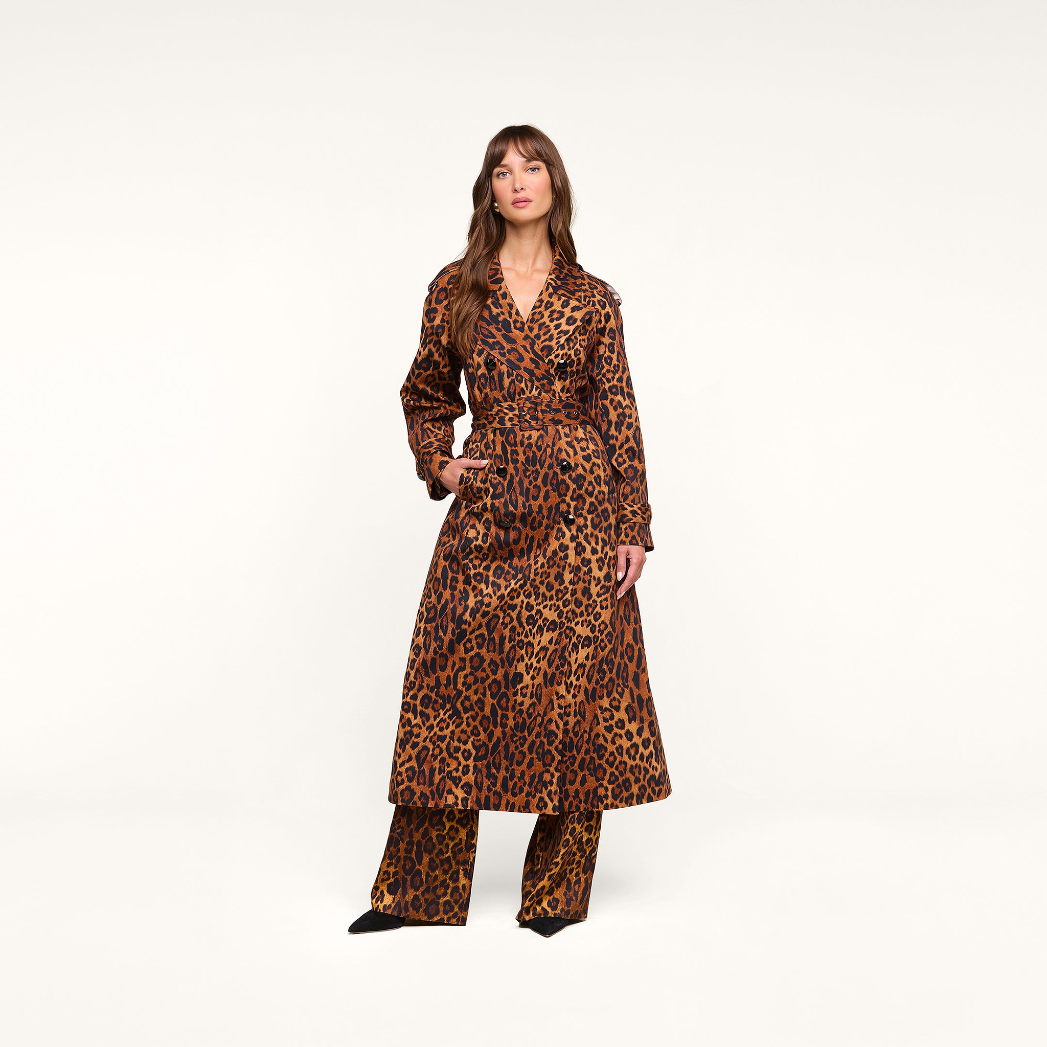 A woman stands against a plain background wearing the Brinley Oversized Trench Coat, a long belted leopard print style, paired with matching leopard print pants and black pointed shoes.