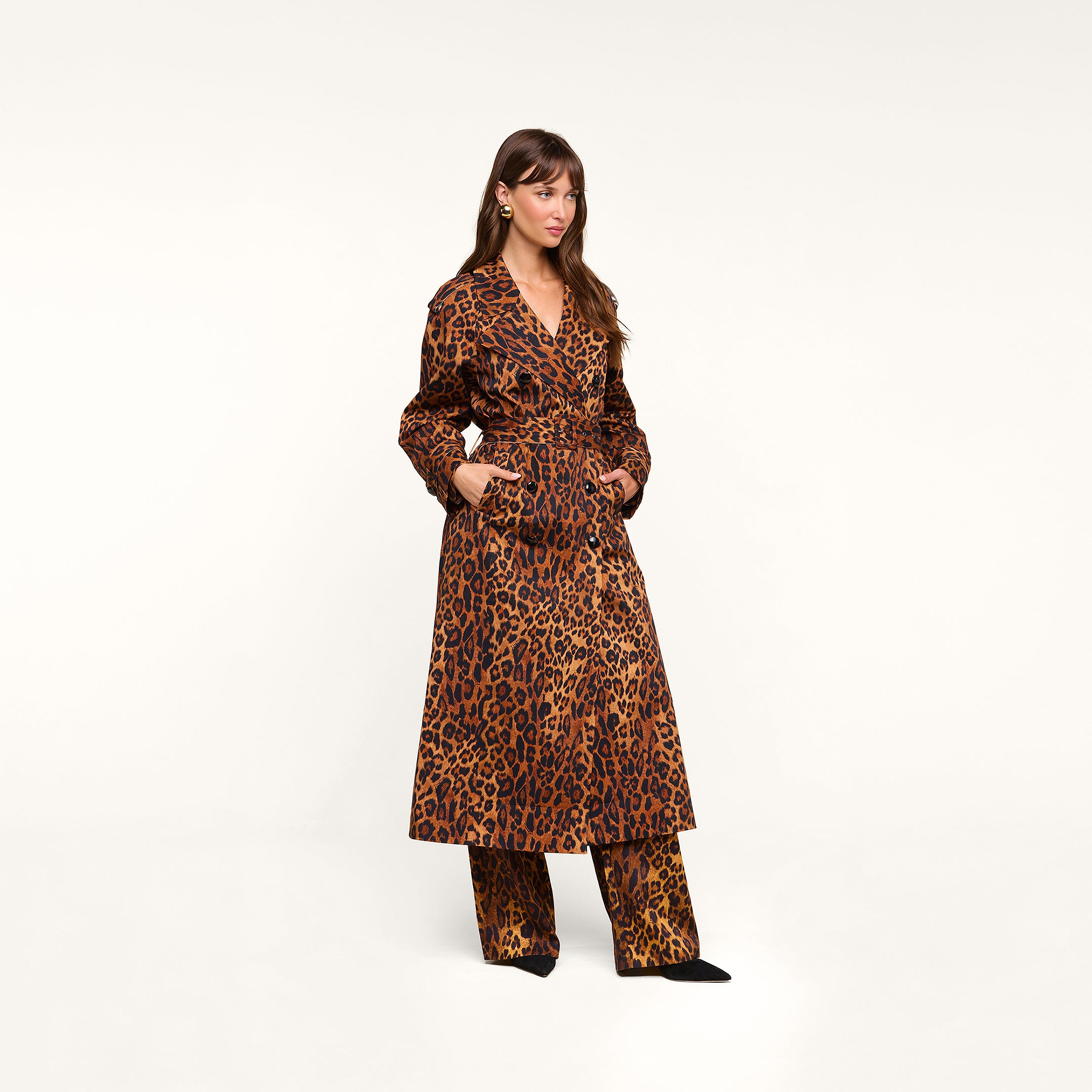 A woman stands against a plain background, wearing the Brinley Oversized Trench Coat in a double-breasted leopard print over matching leopard-print pants. She completes the look with black shoes and gold earrings, her hands tucked into her coat pockets.