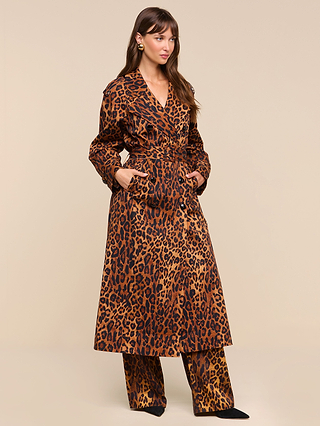 A woman stands against a plain beige background with her hands in the pockets of the Brinley Oversized Trench Coat, styled in a long leopard print and paired with matching wide-leg pants. She wears statement earrings and has long brown hair.