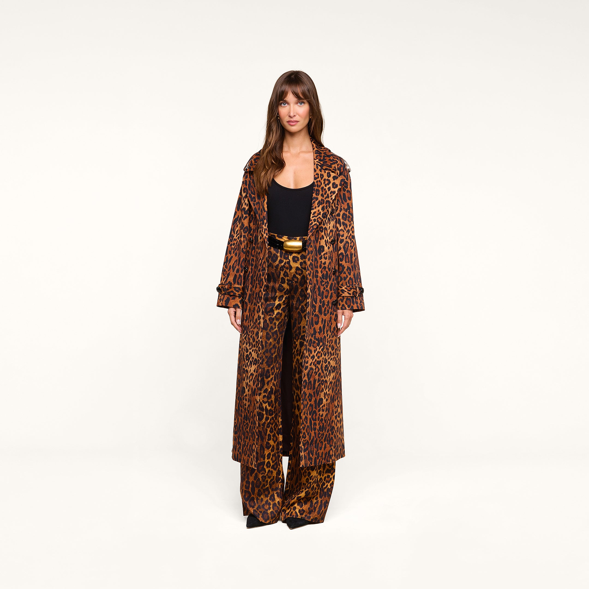 A woman stands facing forward against a plain white background, wearing the Brinley Oversized Trench Coat in a long, double-breasted leopard print, paired with matching wide-leg pants over a black top and accessorized with a gold-buckled belt.