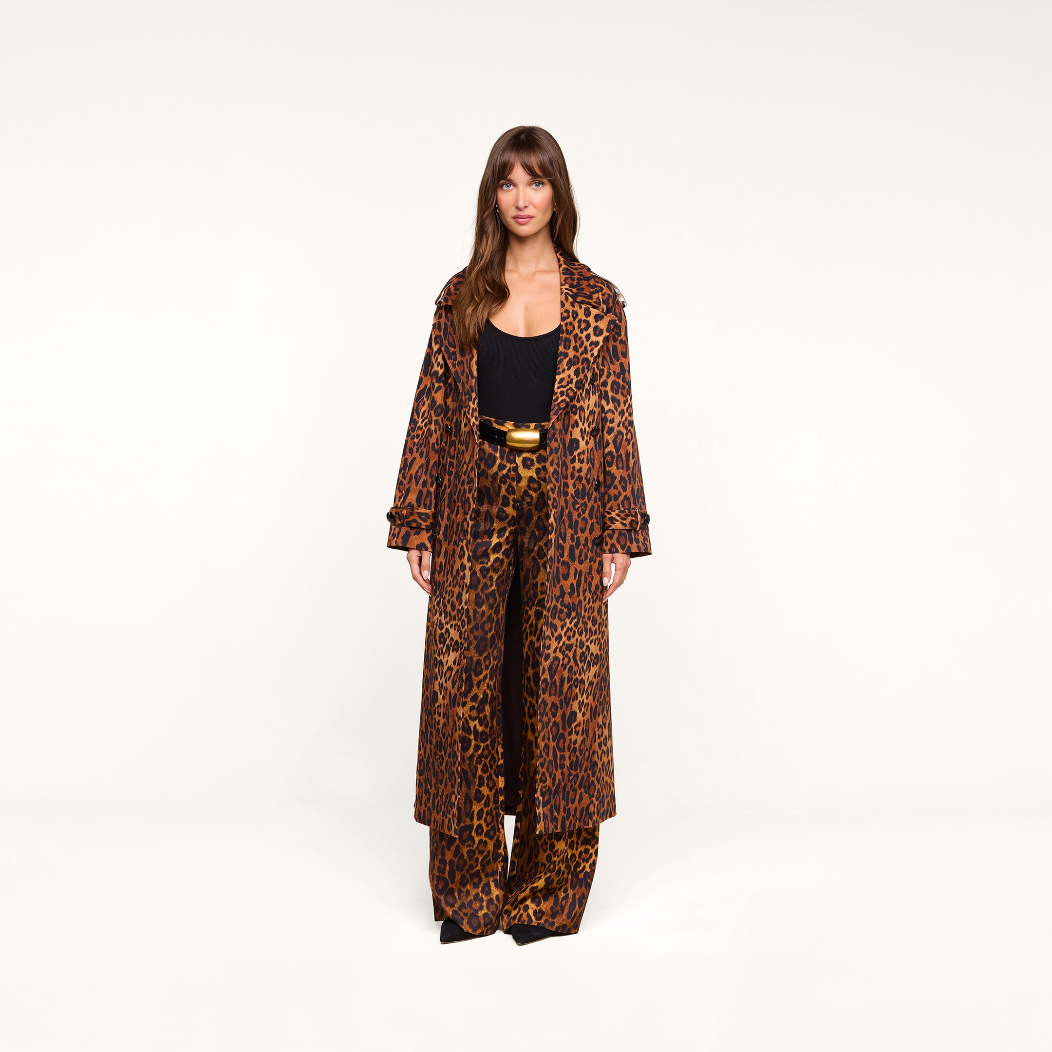 A woman stands facing forward against a plain white background, wearing the Brinley Oversized Trench Coat in a long, double-breasted leopard print, paired with matching wide-leg pants over a black top and accessorized with a gold-buckled belt.
