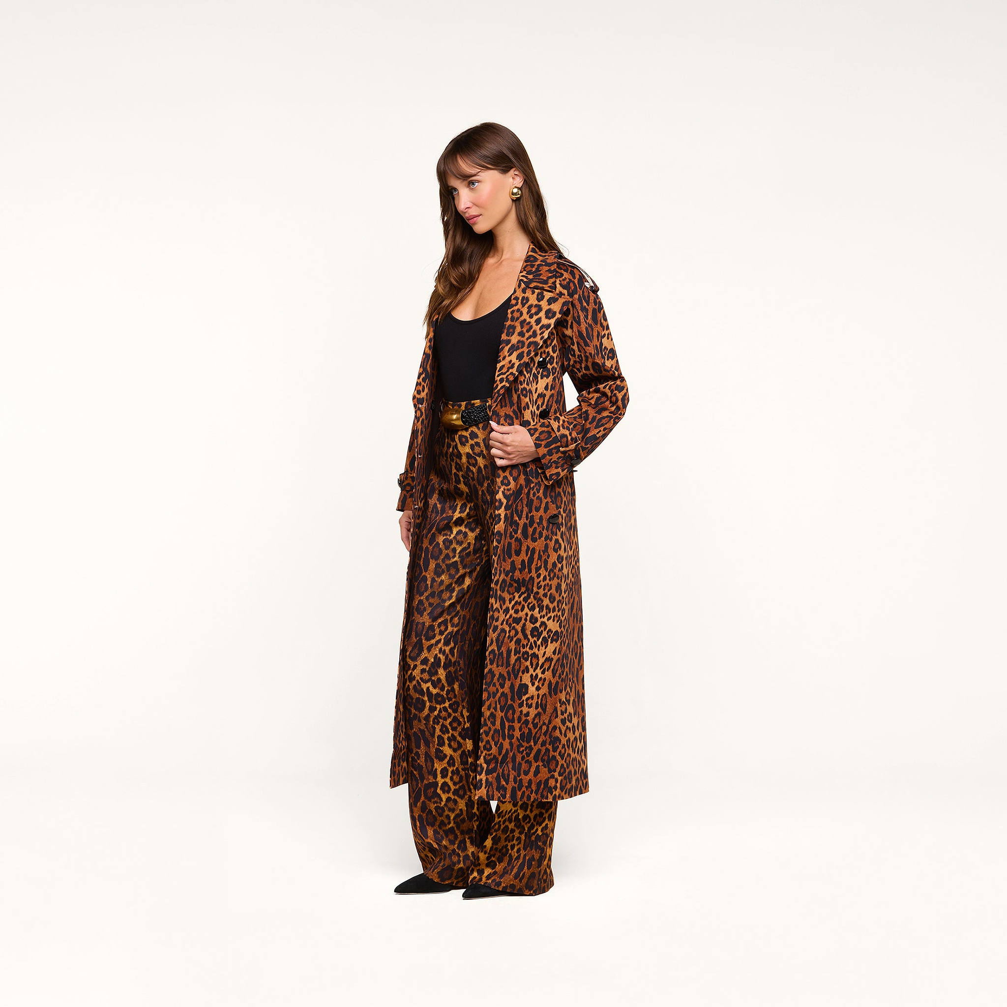 A woman stands against a white background wearing the Brinley Oversized Trench Coat in a double-breasted leopard print, paired with matching leopard-print pants over a black top, her hair down and hands in her coat pockets.
