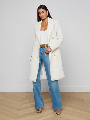 A woman with long brown hair poses indoors against a plain wall, wearing the Cooper Tweed Trench Coat in textured white over a white top, blue high-waisted jeans, brown belt, and open-toe heels on a wooden floor.