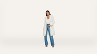 Woman wearing a long white coat, white top, blue wide-leg jeans, and brown heels stands against a plain white background.