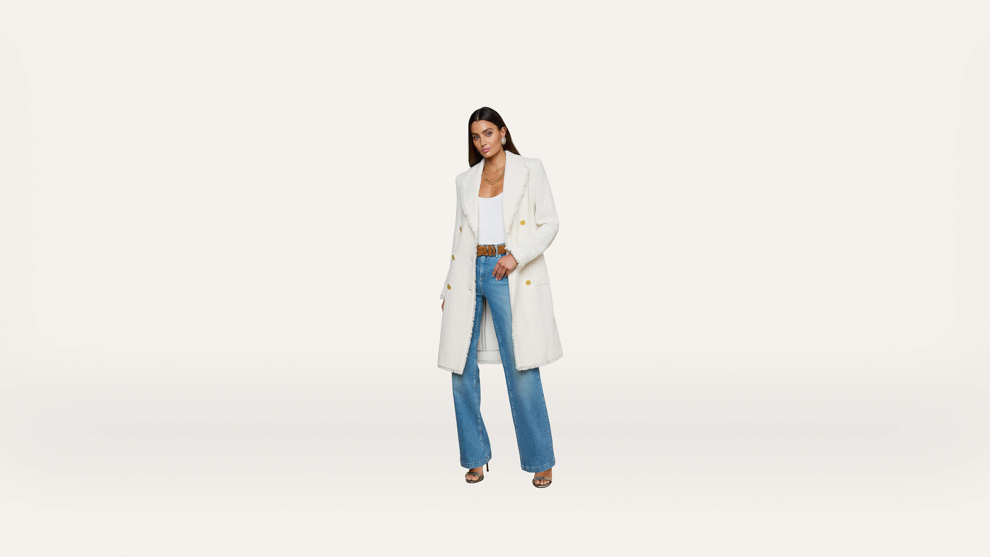 Woman wearing a long white coat, white top, blue wide-leg jeans, and brown heels stands against a plain white background.