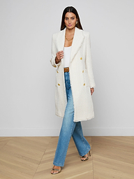 A woman with long brown hair and gold accessories stands on a wooden floor against a white wall, wearing the Cooper Tweed Trench Coat—a long white tweed jacket with frayed trims—over a white top and blue jeans.