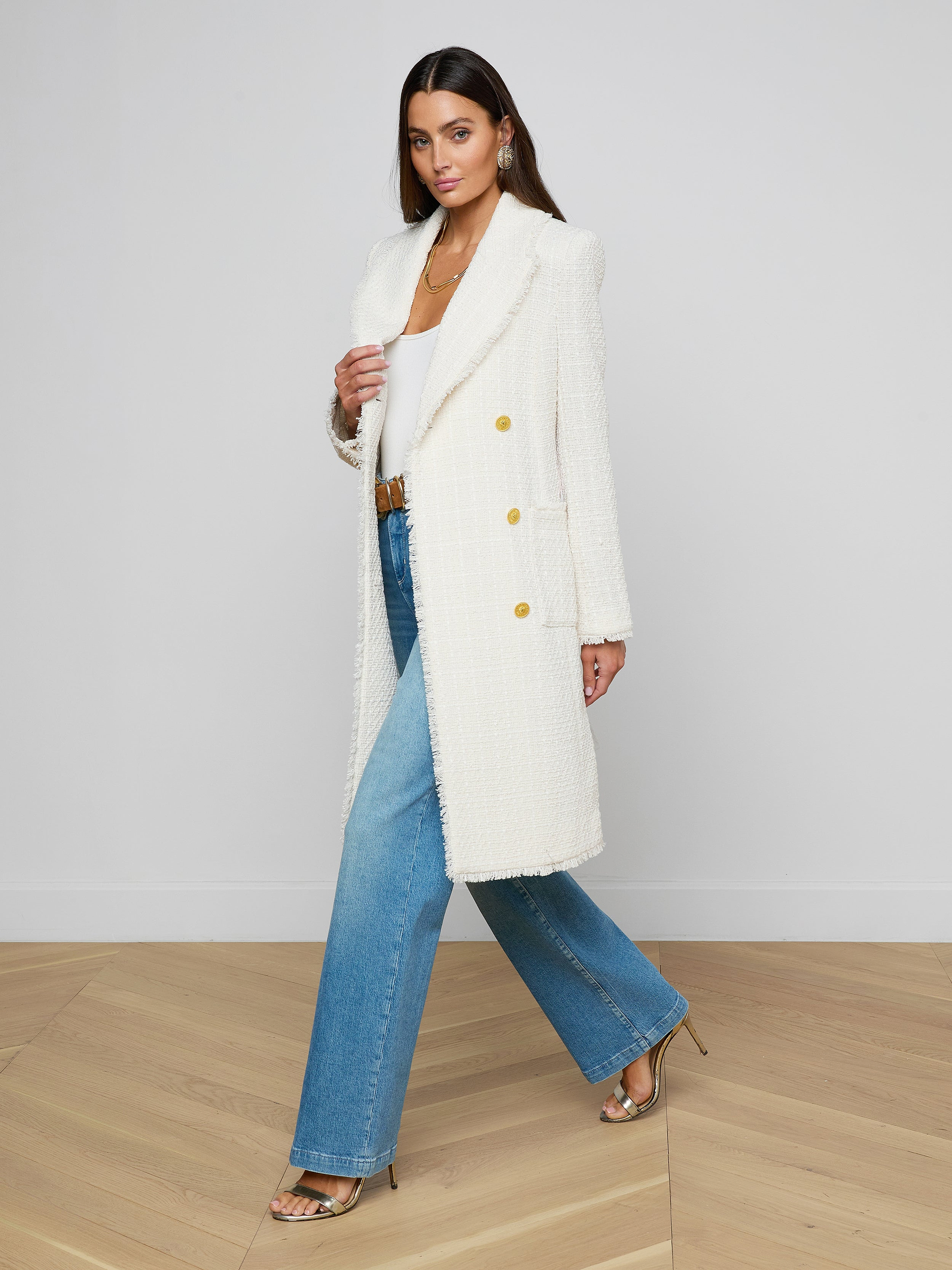 A woman walks confidently on a wooden floor against a white wall, dressed in the Cooper Tweed Trench Coat with frayed trims, a white top, blue wide-leg jeans, and metallic heels.