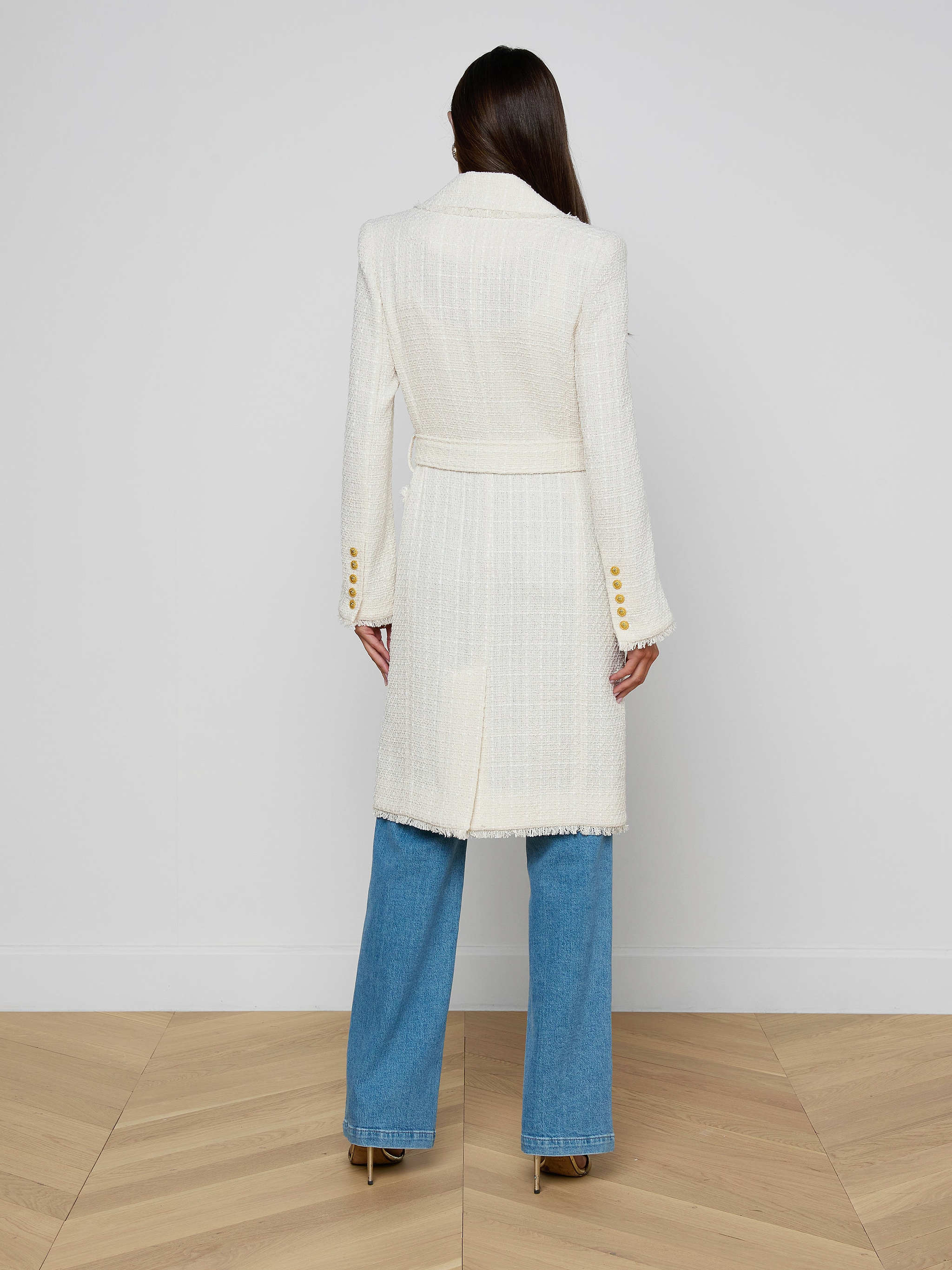 A woman stands facing away on a light wooden floor against a plain white wall, wearing the Cooper Tweed Trench Coat—an unlined long white coat with gold buttons and a belt—paired with blue jeans and heeled shoes.