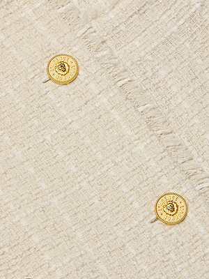 Close-up of the Cooper Tweed Trench Coat's cream unlined tweed fabric with a subtle plaid, featuring frayed trims and two gold decorative buttons embossed with text and a central emblem.