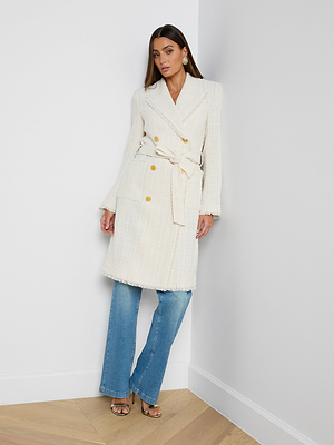 A woman stands in a corner wearing the Cooper Tweed Trench Coat, featuring a textured cream hue and gold buttons, paired with blue jeans, heeled sandals, and statement earrings. Her hair is long and straight, and she has a neutral expression.