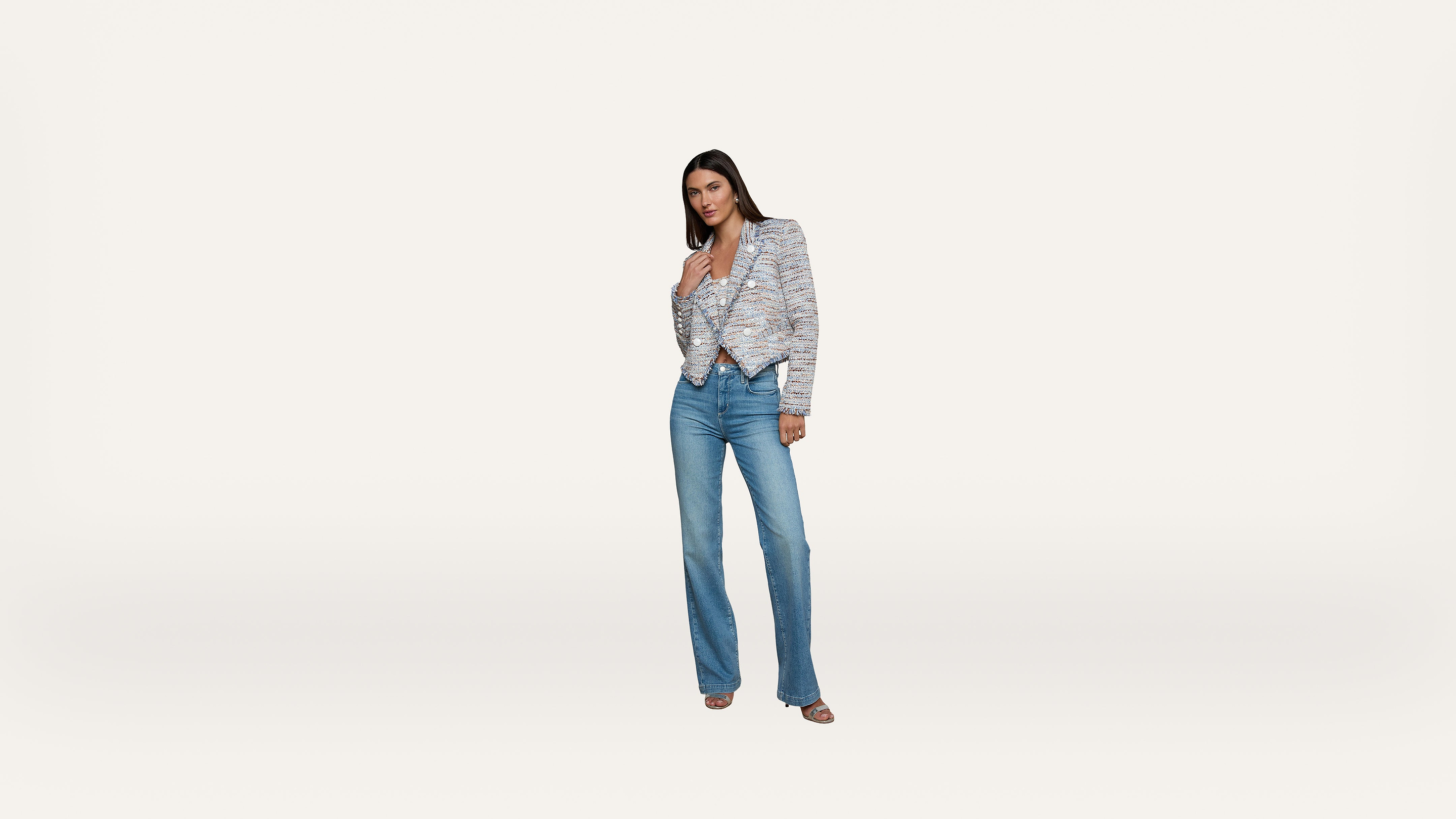 A woman stands against a plain white background, wearing a patterned blazer, a light top, blue jeans, and open-toed heels, with one hand touching her chest and the other by her side.