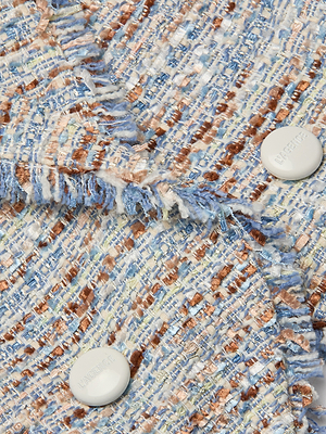 Close-up of the Marv Tweed Blazer's textured blue, white, and brown fabric with frayed edges. Two statement buttons feature faint embossed text, while the woven pattern gives a soft, tactile look.