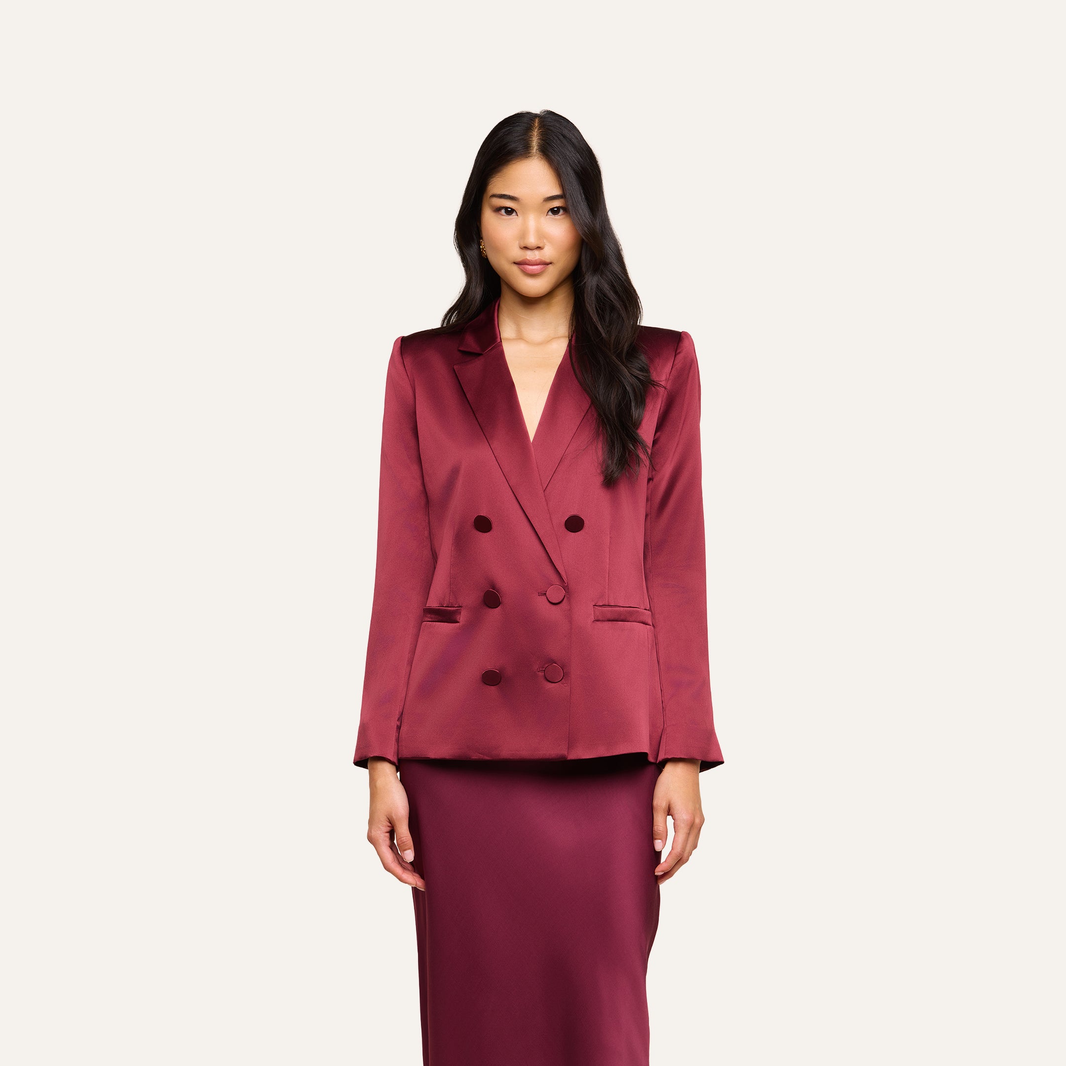 A woman with long black hair wears the Sena Silk Blazer in a shiny merlot red, paired with a matching skirt, standing against a plain light background.