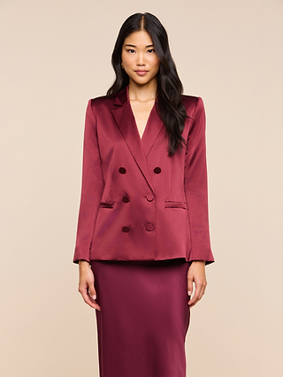 A woman with long dark hair wears the Sena Silk Blazer in merlot red with a matching long skirt, standing against a plain beige background.