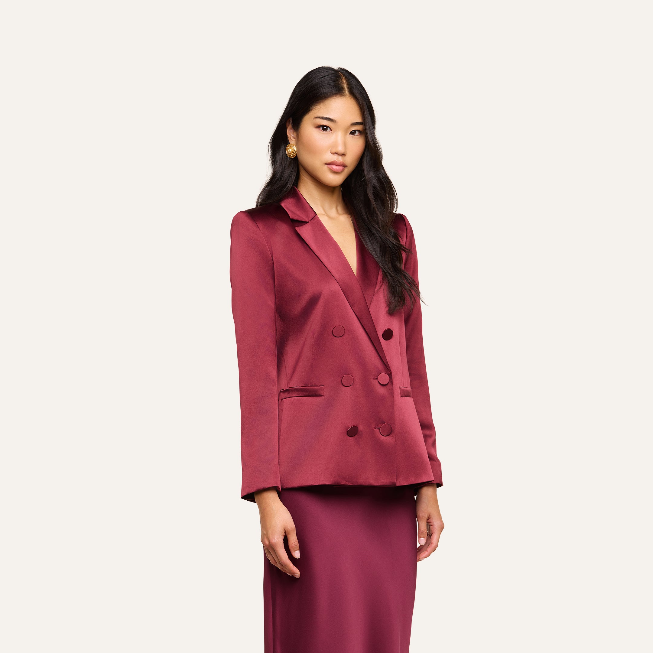 A woman with long dark hair wears the Sena Silk Blazer in merlot red, paired with a matching skirt, standing against a plain light background.