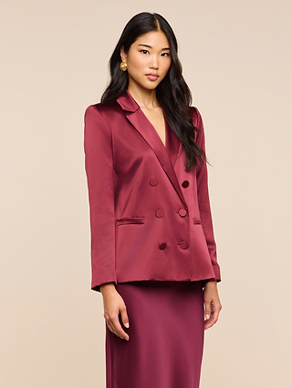 A woman with long dark hair wears the Sena Silk Blazer in merlot red, paired with a matching skirt, standing against a plain beige background. She is facing forward with a neutral expression.