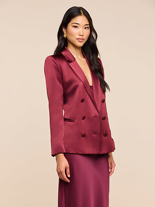 A woman with long dark hair wears the Sena Silk Blazer in shiny merlot red, styled with the matching skirt, standing against a light beige background and looking at the camera with a neutral expression.