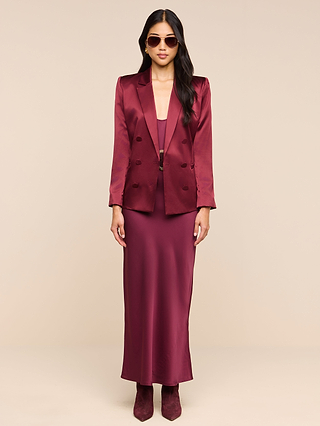 A woman with long dark hair and a confident pose stands against a beige background, wearing the Sena Silk Blazer in merlot red satin, paired with a matching long dress, boots, and sunglasses.