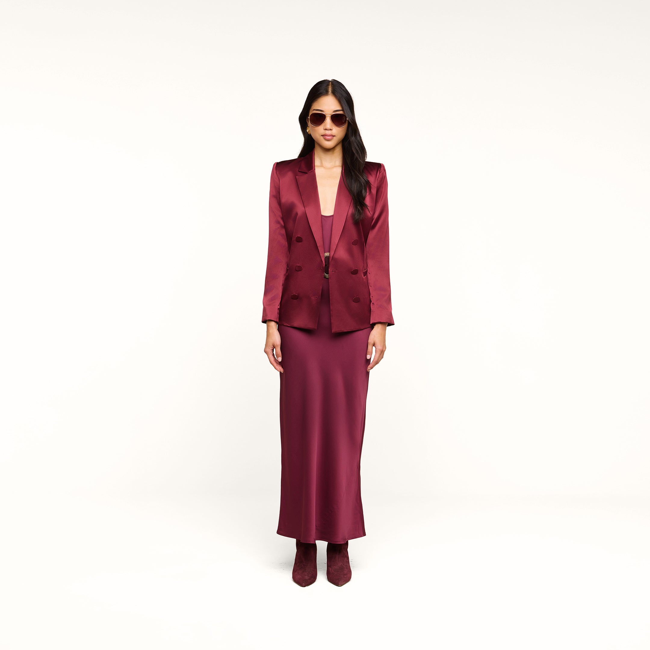 A woman stands against a white background wearing the Sena Silk Blazer in merlot red, paired with a matching long dress, burgundy boots, and sunglasses.