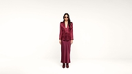 A woman wearing sunglasses, a burgundy blazer, a matching long skirt, and ankle boots stands against a plain white background.