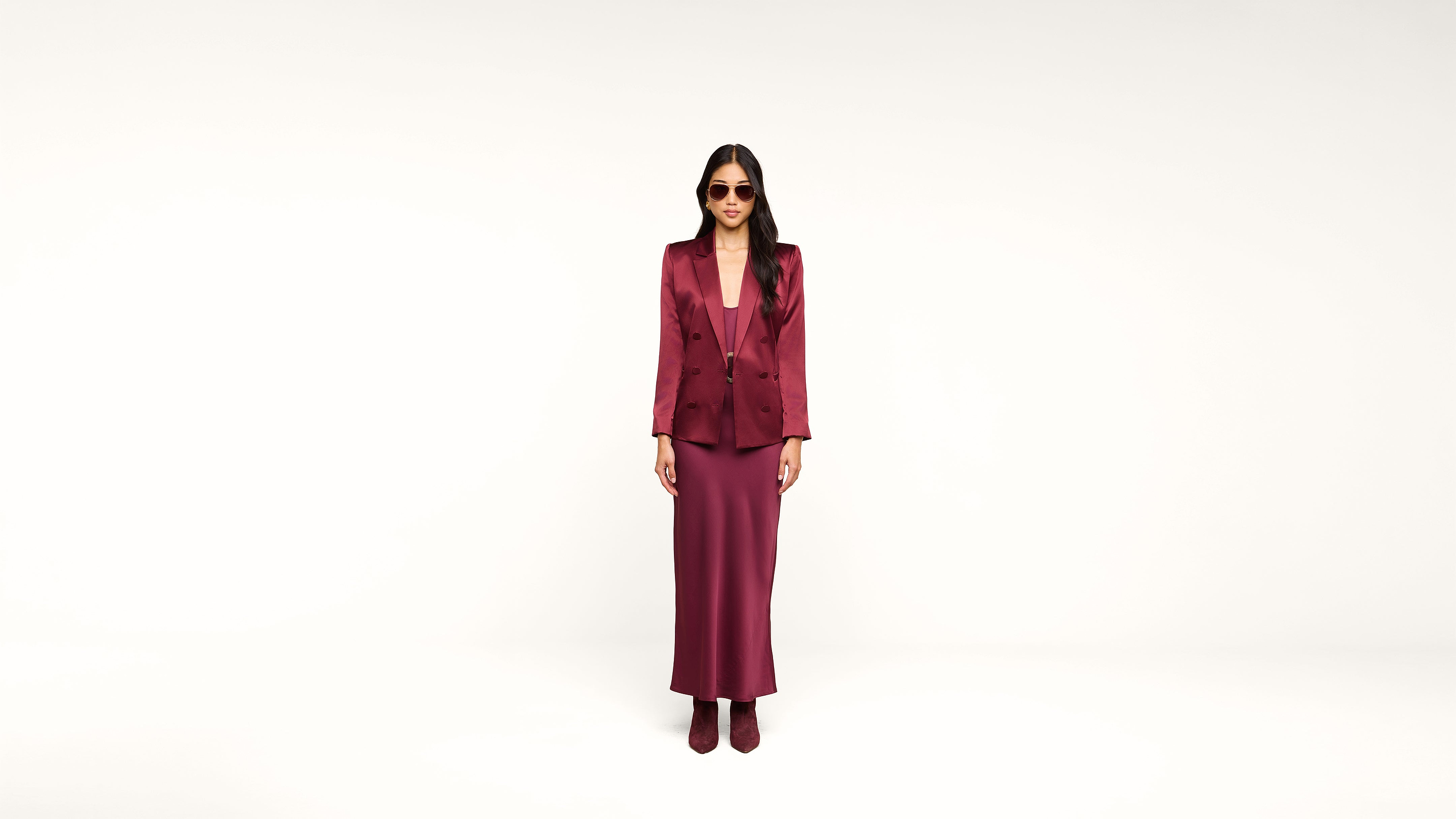 A woman wearing sunglasses, a burgundy blazer, a matching long skirt, and ankle boots stands against a plain white background.