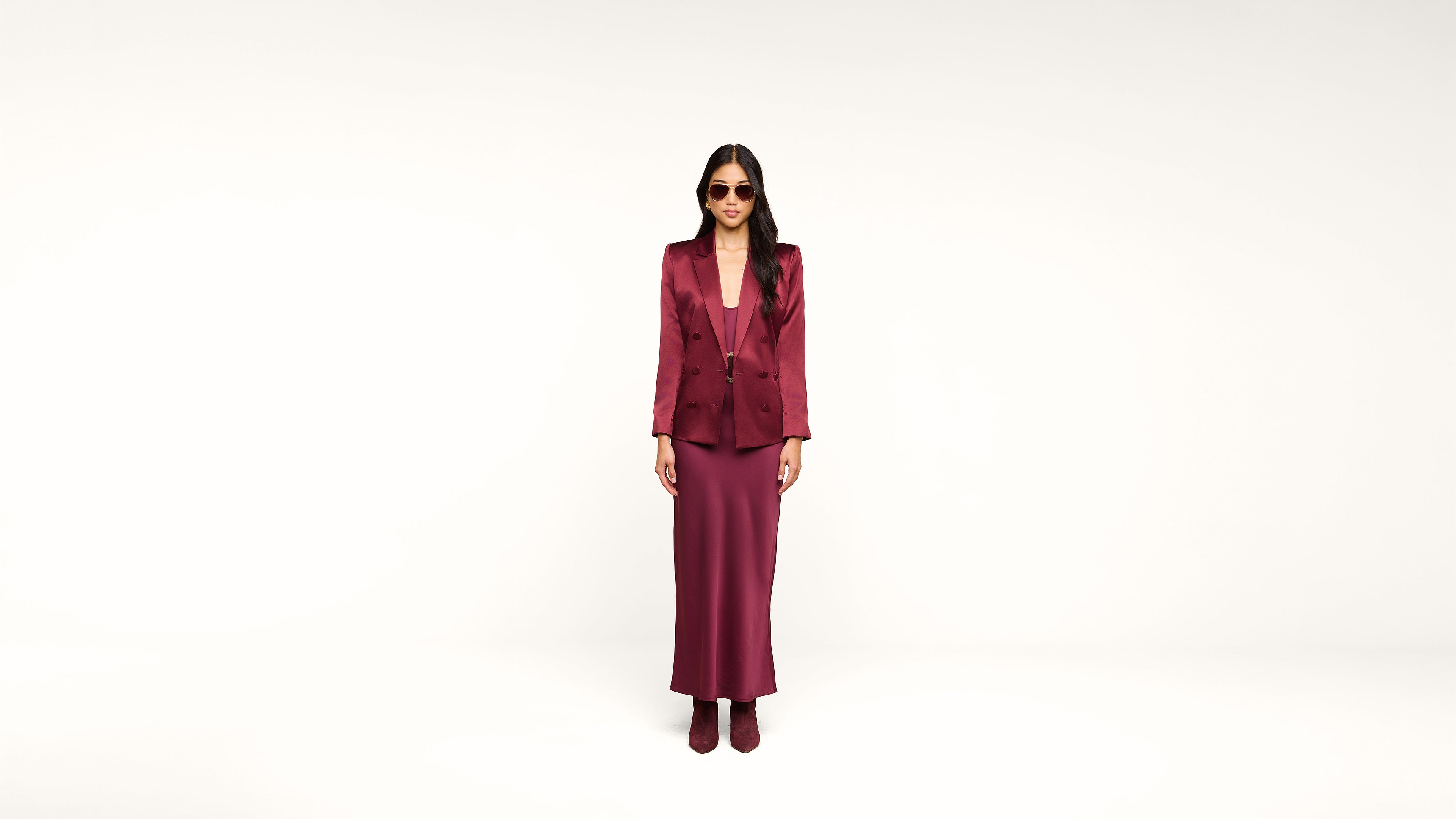 A woman wearing sunglasses, a burgundy blazer, a matching long skirt, and ankle boots stands against a plain white background.