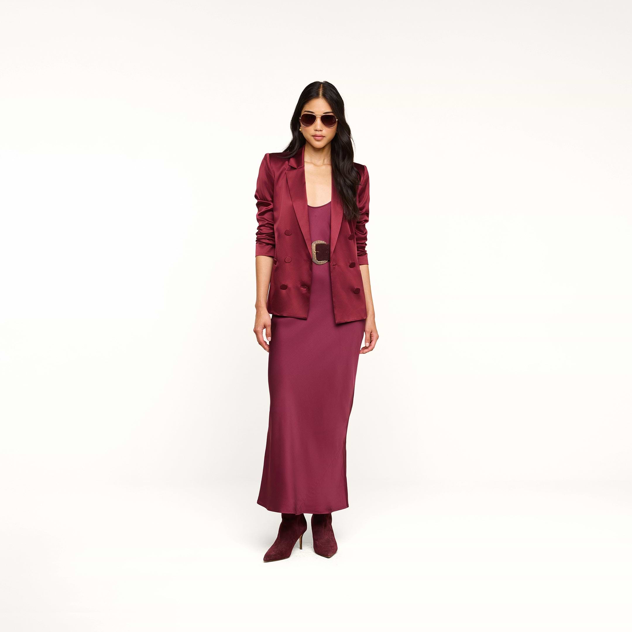 Woman wearing sunglasses, the Sena Silk Blazer in merlot red, a matching dress, and ankle boots standing against a plain white background.