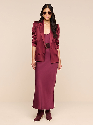 A woman wearing sunglasses and the Sena Silk Blazer in merlot red, styled over a matching dress with a wide belt and paired with maroon ankle boots, stands against a neutral background.