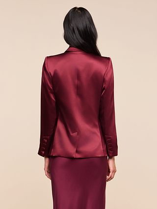 A woman with long dark hair is shown from the back against a plain beige background, wearing the shiny merlot red Sena Silk Blazer with a matching skirt.