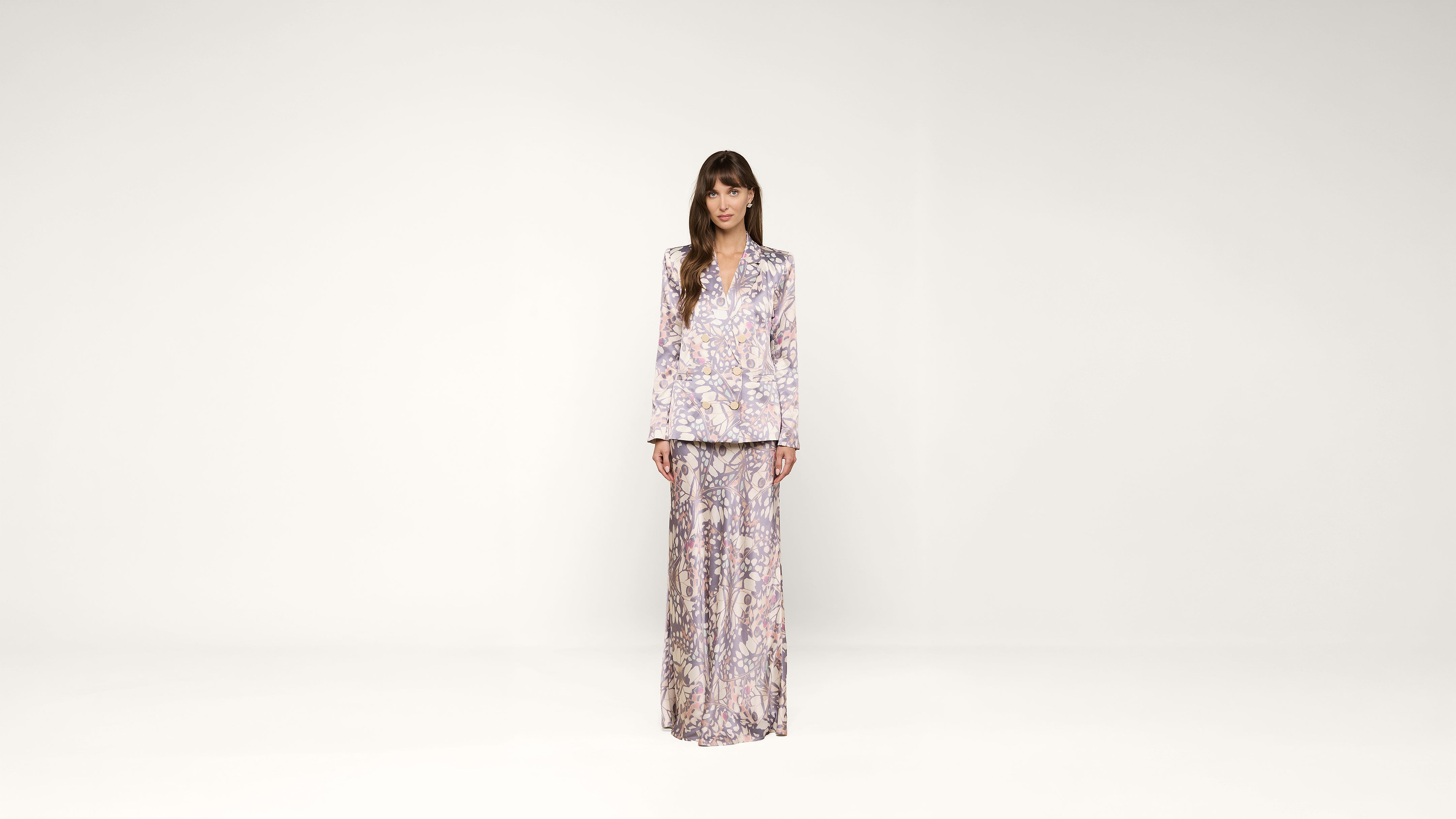 Woman standing against a plain white background, wearing a matching light-colored, floral-patterned blouse and wide-leg pants, with straight brown hair and a neutral expression.