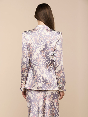A person with long brown hair is seen from behind, wearing the Sena Silk Blazer—a double-breasted set with a pastel abstract floral pattern in purple, pink, and white on a beige background.
