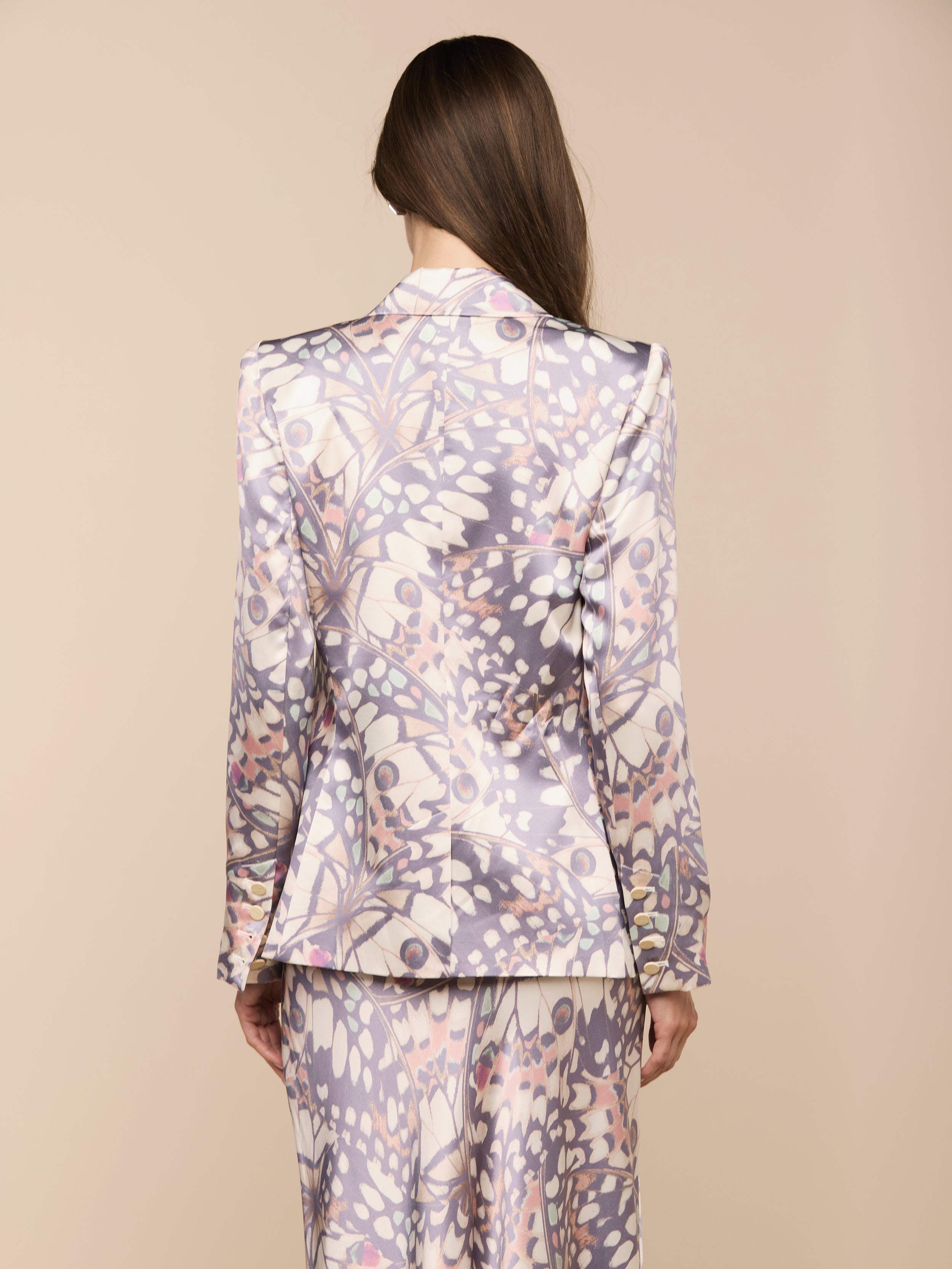 Von hinten sieht man eine Person mit langen braunen Haaren, die den Sena-Seidenblazer trägt - einen Zweireiher mit einem pastellfarbenen abstrakten Blumenmuster in Lila, Rosa und Weiß auf beigefarbenem Hintergrund.