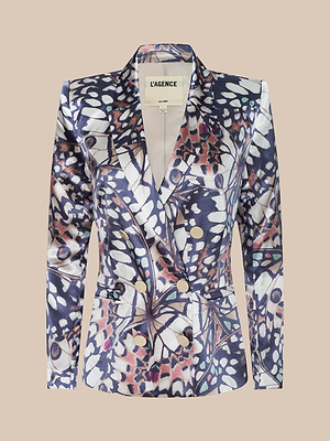 The Sena Silk Blazer is a tailored double-breasted blazer featuring a bold butterfly wing print in navy, white, and beige, accented with gold buttons and displayed against a plain beige background.