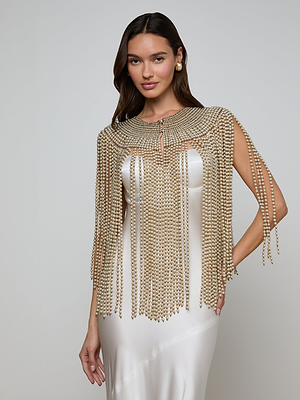 A woman in a white satin dress wears the Yves Cape, featuring long strands and simulated pearls. She stands before a plain light gray backdrop, gazing at the camera with a soft smile that radiates artisanal refinement.