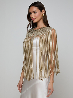 A woman in a white satin dress wears the Yves Cape, featuring long draped strands and matching gold earrings, exuding artisanal refinement against a plain light gray background.