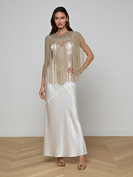 A woman in a white satin floor-length dress, exuding elegance, wears the Yves Cape—a gold beaded fringe capelet. She poses indoors on a wooden herringbone floor with one hand on her hip and the other by her side.