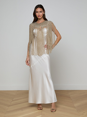 A woman in a white satin floor-length dress, exuding elegance, wears the Yves Cape—a gold beaded fringe capelet. She poses indoors on a wooden herringbone floor with one hand on her hip and the other by her side.