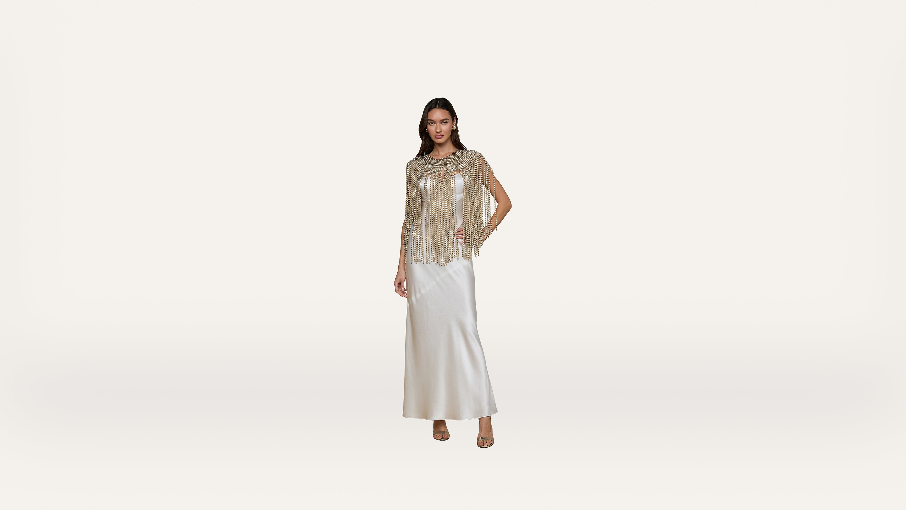 A woman stands against a white background, wearing a long white dress and a sheer, beaded top. She has one hand on her hip and is wearing open-toe heels.