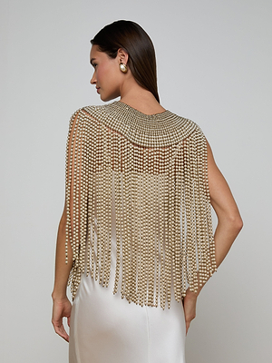 A woman in a cream satin dress is shown from the back, wearing the Yves Cape with gold beaded fringe and simulated pearls. She has long brown hair and gold earrings, standing against a plain light gray background.