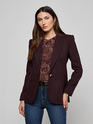 A woman with long brown hair wears the Sheila Longline Blazer over a patterned top and blue jeans. She stands facing the camera with a neutral expression, hand in pocket, against a plain gray background.