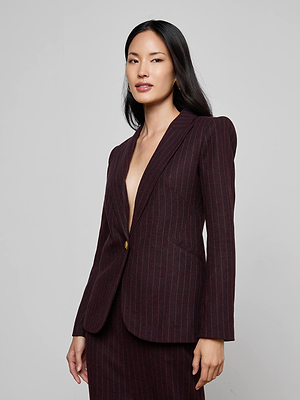 A woman with long dark hair wears the Sheila Longline Blazer—an Italian wool-blend in deep burgundy pinstripes—with no shirt underneath. She stands confidently against a plain light gray background, smiling softly and facing slightly left.